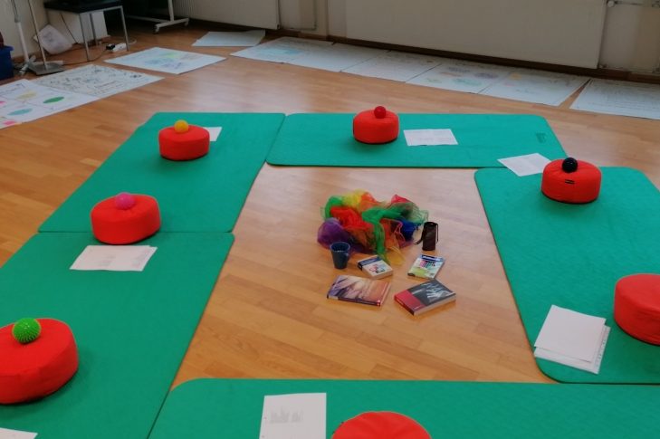 Einlassen auf kreative Wege Orangefarbene Sitzkissen in einem Kreis um Bücher und Materialien auf dem Boden.