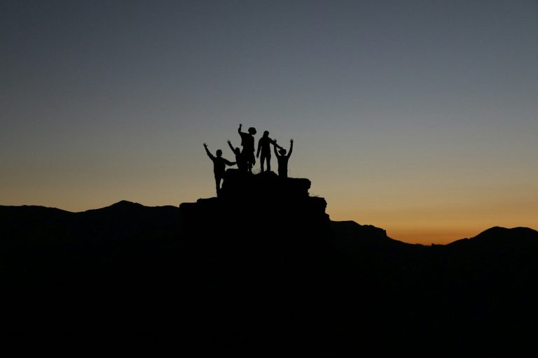 Gemeinsam stark und widerstandsfähig Silhouetten von Personen auf einer Bergspitze. Im Hintergrund der Sonnenuntergangshorizont.